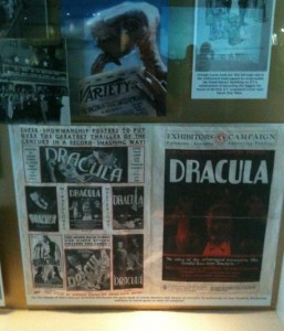 The image shows a display case containing various vintage posters and promotional materials for the movie "Dracula." The display includes a large poster with the title "Dracula" prominently featured, along with smaller images and text detailing the film's promotion. There are also other related images and documents, including a Variety magazine cover and other promotional materials. The display appears to be part of an exhibition or museum exhibit dedicated to the film.