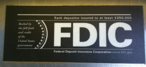 Alt text: A black and white sign with the text "Each depositor insured to at least $250,000" at the top. Below this, in large letters, is "FDIC." To the left of "FDIC" is a circular emblem with stars and the text "Backed by the full faith and credit of the United States government." At the bottom, it reads "Federal Deposit Insurance Corporation - www.fdic.gov."