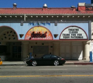 The image shows the exterior of a closed theater with a sign that reads, "THEATRE CLOSED THANK YOU FOR YOUR PATRONAG." The building has a decorative facade with an arched design and a marquee. There is a black car parked in front of the theater, and a bicycle is also visible nearby. The theater's name appears to be partially visible above the marquee.