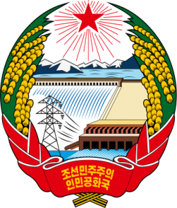 The image is the national emblem of North Korea. It features a hydroelectric dam with water flowing from it, a transmission tower, and a power plant. Surrounding the central image are sheaves of rice, and above it is a red star with rays emanating from it. At the bottom, there is a red ribbon with Korean text.