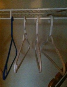 The image shows a close-up of a closet rod with four empty clothes hangers hanging on it. Three of the hangers are white, and one is blue. The background is dimly lit.