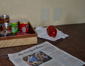 The image shows a countertop with a few items on it. There is a wicker basket containing cans of nuts and two cans of Pringles chips. Next to the basket, there is a red bell pepper placed on a piece of white paper. In the foreground, there is a copy of The New York Times newspaper. The background features a beige wall with two electrical outlets.