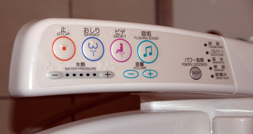 The image shows the control panel of a bidet toilet seat. The panel includes various buttons with icons and text in both Japanese and English. The buttons are labeled for functions such as "STOP," "SPRAY," "BIDET," "FLUSHING SOUND," "WATER PRESSURE," and "VOLUME." There are also buttons for power and other settings. The panel is white with colored icons and text.