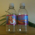 The image shows two plastic water bottles placed side by side on a wooden surface. Each bottle has a label with a pink border. The label on the left bottle features an image of a woman in a white top and the text "CALIF V" and "PURIFIED DRINKING WATER." The label on the right bottle has the text "CALENDAR GIRL WATER" and "HOOTERS" with an image of a woman's legs. There are green plant leaves in the background.