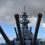 The image shows the front view of a battleship with several large naval guns pointing forward. The ship's superstructure, including radar and communication equipment, is visible in the background. The sky is partly cloudy with patches of blue. The ship appears to be docked at a port, with some buildings and other structures visible in the distance.