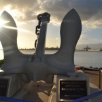 The image shows a large anchor monument dedicated to the USS Arizona, with a plaque commemorating the date December 7, 1941, and the phrase "Remember, Understand, Honor." The monument is situated by the water, with a scenic view of the harbor and a bridge in the background. The sun is setting or rising, casting a warm glow over the scene.