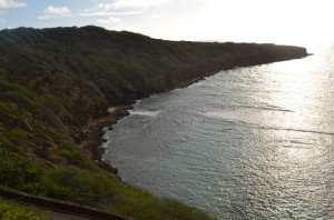 The image shows a coastal landscape with a large body of water bordered by a rugged, green hillside. The sun is shining brightly, creating a reflection on the water's surface. The coastline curves gently, and the sky above is partly cloudy. The overall scene appears serene and natural.