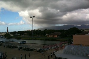 The image shows a cloudy sky over a landscape with a mix of buildings, trees, and a body of water. There is a tall light pole in the middle of the scene, and a few vehicles and people are visible on the ground. The background includes mountains partially obscured by clouds. The overall atmosphere appears to be overcast with a potential for rain.