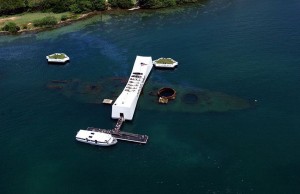 Aerial view of the USS Arizona Memorial at Pearl Harbor, Hawaii. The white structure of the memorial spans the sunken remains of the battleship USS Arizona, which is visible beneath the water. A boat is docked at the memorial, and there are small green islands in the background. The water is a deep blue-green color.