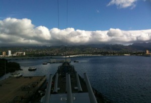 The image shows a view from the deck of a battleship looking out over a harbor. The harbor is surrounded by land with buildings and mountains in the background. The sky is partly cloudy with a mix of white clouds and blue sky. Several boats are visible in the water near the shore.