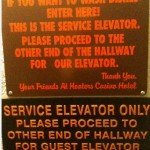 The image shows two signs with text. The top sign reads: "IF YOU WANT TO WASH DISHES ENTER HERE! THIS IS THE SERVICE ELEVATOR. PLEASE PROCEED TO THE OTHER END OF THE HALLWAY FOR OUR ELEVATOR. Thank you, Your Friends At Hooters Casino Hotel." The bottom sign reads: "SERVICE ELEVATOR ONLY PLEASE PROCEED TO OTHER END OF HALLWAY FOR GUEST ELEVATOR." Both signs have orange text on a dark background.