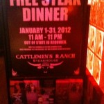 The image shows a promotional sign for a "Free Steak Dinner" at Cattlemen's Ranch Steakhouse. The offer is valid from January 1-31, 2012, between 11 AM and 11 PM. The sign specifies that an out-of-state ID is required and that the offer is limited to one per customer per visit. The sign also mentions that the purchase of a beverage is required for the free steak dinner. The background of the sign is dark, and it is illuminated by a warm, reddish light.