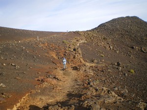 A person is walking on a rocky, barren trail in a mountainous area. The landscape is mostly brown and dry, with scattered rocks and sparse vegetation. The sky is clear with a few clouds.