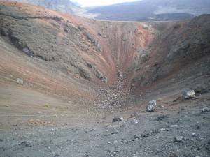 The image shows a barren, rocky crater with steep sides. The ground is composed of loose, dark volcanic rock and soil, with some larger rocks scattered around. The crater appears to be in a remote, mountainous area with a desolate landscape. The sky is overcast, adding to the stark and rugged appearance of the scene.