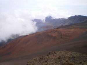 The image shows a mountainous landscape with a mix of reddish and brown terrain. There are clouds partially covering the mountains in the background, and the sky is mostly overcast. The scene appears to be taken from a high vantage point, providing a wide view of the rugged and barren landscape.