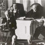 A black and white photograph of a formal event, likely a political or governmental address. A man is standing at a podium with multiple microphones, speaking to an audience. Behind him, several other men are seated, some at a higher elevation, suggesting a position of authority or importance. The setting appears to be an official chamber or hall, with a large desk and papers in front of the seated men. The atmosphere is serious and formal.