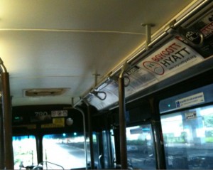 The image shows the interior of a bus. The view is from the back, looking towards the front of the bus. There are handrails and hand straps hanging from the ceiling for passengers to hold onto. There are advertisements displayed above the windows on the right side. The bus appears to be in motion, as the view through the front windshield shows a blurred scene of the road and surroundings. The lighting inside the bus is dim.