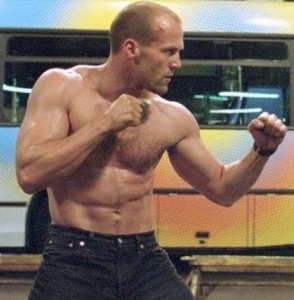 A shirtless, muscular man is standing in a fighting stance with his fists raised. He is outdoors, and there is a bus with a colorful design in the background.