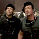 Two men are standing side by side, both wearing black tactical gear and black berets. They appear to be in a serious or contemplative mood, looking slightly upwards. The background is blurred but seems to depict a rugged or chaotic environment, possibly with debris or equipment.