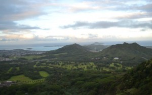 The image shows a scenic landscape with rolling green hills and mountains under a partly cloudy sky. In the distance, there is a body of water, possibly a lake or the ocean, and some scattered buildings or structures. The foreground is lush with greenery, including trees and grassy areas. The overall scene is serene and natural.