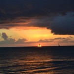 A serene ocean scene at sunset with a dramatic sky. The sun is partially hidden by clouds, casting a warm, golden glow across the water. Two sailboats are visible on the horizon, silhouetted against the vibrant colors of the sunset. Dark clouds loom above, adding contrast to the bright hues of the setting sun.