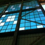 The image shows a large industrial window composed of multiple square panes of glass. The panes are primarily blue, with a few white ones interspersed. The window is framed by a metal grid structure, and some of the panes appear to be cracked or damaged. The view is from inside a building, looking up towards the window.