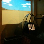 The image shows a flight simulator setup. There is a cockpit with various controls and instruments, and a large screen in front of it displaying a sky with clouds and a landscape, simulating the view a pilot would see. The cockpit appears to be enclosed, and the overall lighting is dim, suggesting an indoor environment.