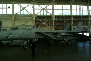 The image shows a military jet aircraft, specifically an F-14 Tomcat, parked inside a large hangar. The aircraft is painted in a gray color and has the number "102" marked on its nose. A person is standing in front of the jet, facing away from the camera. The hangar has a high ceiling with large windows and a metal framework.