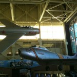 The image shows two vintage military aircraft displayed indoors in a museum setting. The aircraft on the left is a silver jet with red stars on its wings and the number "1524" painted on its fuselage. The aircraft on the right is also silver with a checkered pattern on its tail and the number "12832" on its tail fin. The museum has large windows and a high ceiling with visible structural beams. There are informational displays and signs below the aircraft, and a few people are visible in the background.