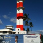 The image shows a tall control tower with red and white horizontal stripes. In front of the tower, there is a sign that reads "Ford Island Control Tower Stabilization Project." The sign provides details about the project, including the start date (January 20, 2011), estimated completion date (November 2012), duration (22 months), and funds needed ($5.5 million). The background includes a clear blue sky with some clouds and several palm trees.