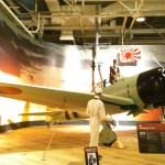 The image shows a museum exhibit featuring a Japanese World War II-era fighter aircraft, likely a Mitsubishi A6M Zero. The aircraft is displayed with a mannequin dressed in a pilot's uniform sitting in the cockpit and another mannequin in a white uniform standing beside the plane. The backdrop includes a large mural with a sunset and a Japanese naval ensign flag. There are informational plaques and a "Keep Off" sign in front of the exhibit. The setting appears to be indoors with visible ceiling structures and lighting.