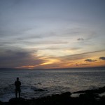 A person stands on a rocky shoreline, gazing out at the ocean during sunset. The sky is filled with a mix of dark and light clouds, with the sun casting a warm, golden glow on the horizon. The water appears calm, and the overall scene is serene and peaceful.