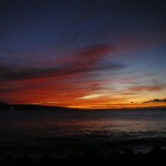 A stunning sunset over the ocean with a vibrant sky displaying shades of red, orange, and purple. The dark silhouette of a distant landmass is visible on the horizon, and the calm water reflects the colors of the sky. The foreground is dark, with the outline of rocks or a shoreline barely visible.