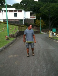 A man is walking on a paved path in an outdoor setting. He is wearing a gray t-shirt, shorts, and sneakers. The background features a white building, green lawns, and trees. There are also some small decorative items or plants along the grassy area to the left of the path. The sky appears to be overcast.