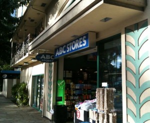 The image shows the exterior of an ABC Stores location. The store has a blue and white sign with the store's name above the entrance. The entrance is open, revealing shelves stocked with various products inside. Outside the store, there are some items on display, including what appears to be a rack of woven mats or rugs. The building has a decorative pattern on the walls near the entrance, and there is a sidewalk in front of the store. The store is located in a building with multiple levels, and there are balconies visible on the upper floors. Trees and other greenery are also visible along the sidewalk.