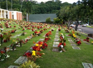 The image shows a well-maintained cemetery with rows of graves adorned with colorful flower arrangements. The graves are marked with flat headstones, and the cemetery is surrounded by lush greenery and trees. There is a building visible on the left side and a road with parked cars on the right side. The overall atmosphere is serene and peaceful.