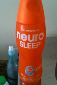 The image shows an orange bottle of "neuro SLEEP" drink. The bottle is labeled as non-carbonated and features the text "ZZZzzz in every bottle" and "No artificial colors or flavors." The bottle is placed on a surface, and there is a dish soap bottle and another container partially visible in the background.