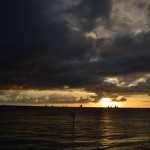 A dramatic sunset over the ocean with dark, heavy clouds partially obscuring the sky. The sun is low on the horizon, casting a golden glow across the water. Numerous sailboats are silhouetted against the horizon, and a person on a paddleboard is visible in the foreground, adding a sense of scale and tranquility to the scene.