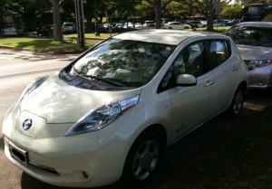 The image shows a white Nissan Leaf electric car parked on the side of a street. The car is positioned in a shaded area with trees and other parked cars visible in the background. The front and side of the car are clearly visible.