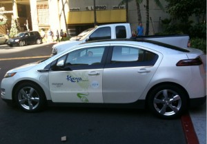 The image shows a white sedan parked on the side of a street. The car has a logo and text on the side that reads "Kona" and "Electric." There is a white pickup truck parked in front of the sedan. In the background, there are buildings, a few people walking, and some trees.