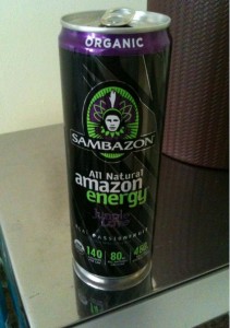 The image shows a can of Sambazon All Natural Amazon Energy drink. The can is predominantly black with purple and green accents. The top of the can is silver with a purple rim that has the word "ORGANIC" written on it. The front of the can features the Sambazon logo, which includes an image of a person wearing a headdress. The text on the can indicates that it is a "Jungle Love" flavor and highlights that it contains 140 calories, 80 mg of caffeine, and 4600 mg of antioxidants. The can is placed on a reflective surface.