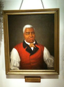 A framed portrait painting of a man with short white hair, wearing a red vest over a white shirt with puffy sleeves, a black collar, and a yellow bow tie. The portrait is hanging on a wall, and there is a small plaque beneath the frame.