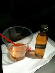 A small bottle of Johnnie Walker Black Label whiskey is placed next to a plastic cup containing a drink with ice and a red straw. The items are on a white napkin.