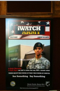 The image is a poster for the "iWATCH ARMY" program. The poster features a soldier in uniform, wearing a beret, standing in front of a building. The top of the poster has the text "iWATCH ARMY" with a background of stars and stripes. Below the title, it says "REPORT" and "KEEP US SAFE." The main text on the poster reads: "1 Simple Observation 1 Simple Report can lead to actions that may STOP a terrorist attack. THINK ABOUT THE POWER OF THAT. THE POWER OF iWATCH. See Something Say Something." At the bottom, there is a logo and the text "Always Ready, Always Alert" along with a website URL.