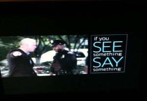 The image shows two police officers standing and talking to each other on the left side. On the right side, there is a message that reads, "If you SEE something SAY something." The background appears to be outdoors with trees and a vehicle visible.