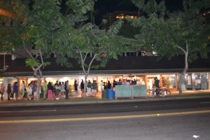 A nighttime scene of a building with a long line of people waiting outside. The building is illuminated, and there are trees in front of it. The street in the foreground is empty, and there are a few bicycles parked near the building.