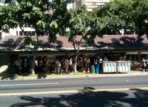 Alt text: A group of people standing in line outside a building on a sunny day. The building has a brown roof and is partially shaded by trees. The street in front of the building has a yellow line marking and is also shaded by tree shadows.