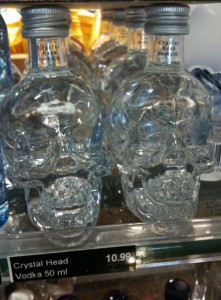 The image shows several small, clear glass bottles shaped like human skulls. They are displayed on a store shelf. The label on the shelf reads "Crystal Head Vodka 50 ml" with a price of "10.99".