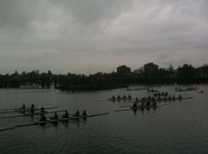 The image shows several rowing teams on a body of water, likely participating in a race or practice session. The sky is overcast, and there are buildings and trees visible in the background. The rowers are in long, narrow boats, and the scene appears to be taking place in an urban area.