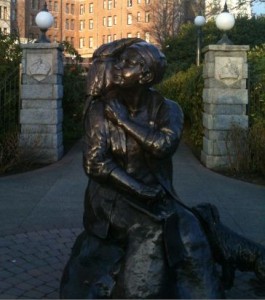 A bronze statue of a woman holding a child is situated in an outdoor setting. The woman is seated and appears to be embracing the child, who is perched on her shoulder. The statue is placed on a stone base, and behind it, there are two stone pillars with spherical lamps on top. In the background, there is a building with multiple windows and some greenery. The scene is captured in daylight.